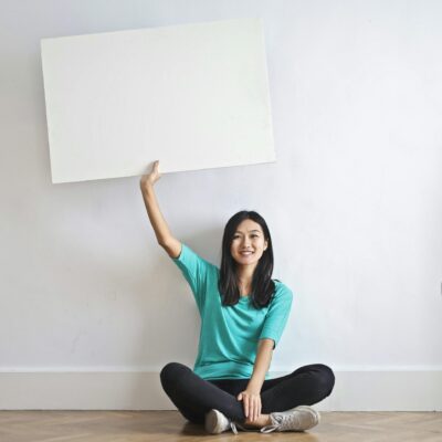 Cheerful Asian woman sitting cross legged on floor against white wall in empty apartment and showing white blank banner