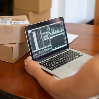 Person Looking at Charts Displayed on a Laptop and Sitting at the Table with Cardboard Boxes