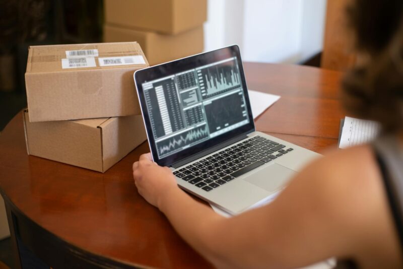 Person Looking at Charts Displayed on a Laptop and Sitting at the Table with Cardboard Boxes